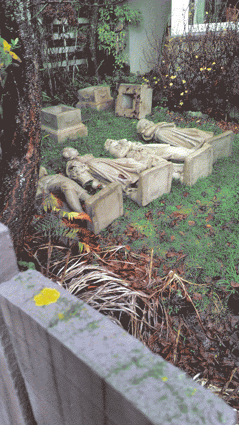 a clandestine photo of four statues lying in someone's front yard. they are laid in a line in front of their pedestals. They look wealthy but not necessarily royal, and seem to be modeled on classical fashion.