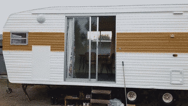 Left hand side of the caravan. Double door in the middle with a table and charis visible. small window to the left near the edge of the caravan. you can see the toilet cassette hatch at the bottom right. sky is overcast, mustard strip very prominent against the white caravan
