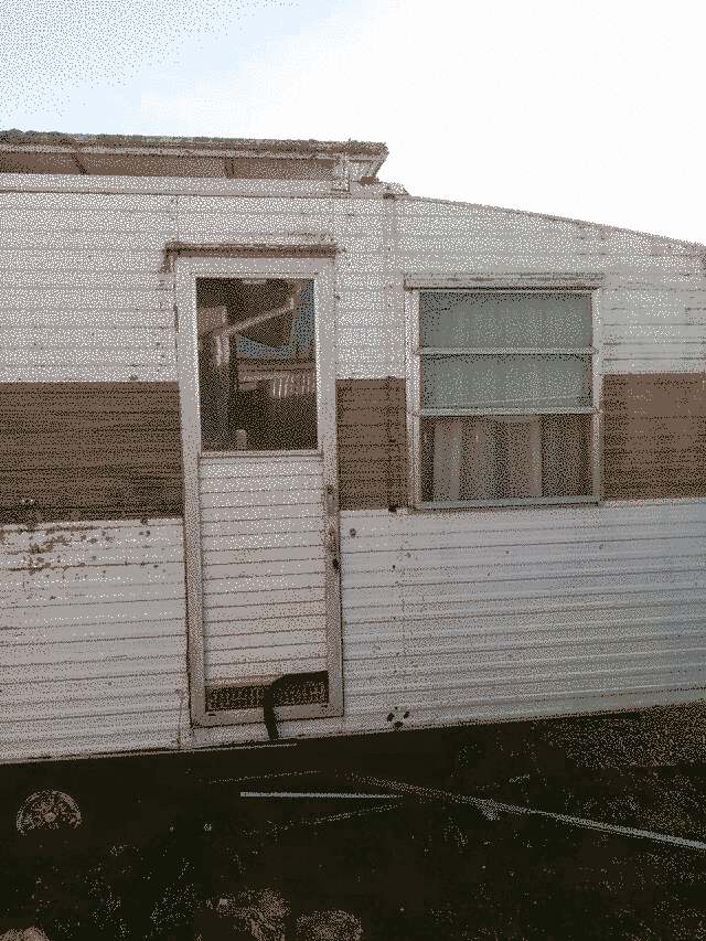 right half of a 1976-ish Millard caravan. grimy, a window and a door, white corrugate with a brown band across the middle
